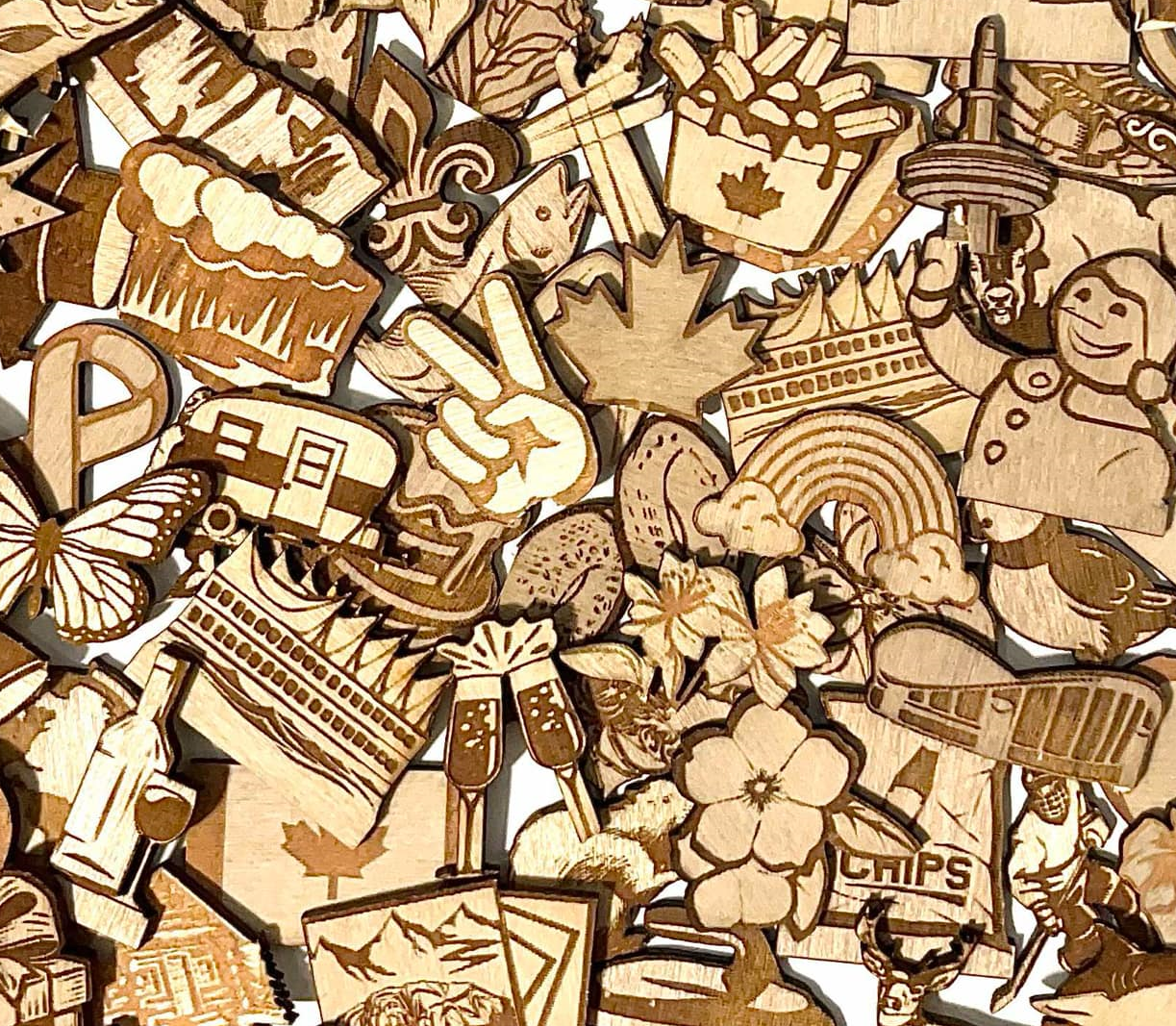 A dense, flat-lay collage of various laser-cut wooden ornaments and decorations, featuring Canadian-themed motifs like maple leaves, a rainbow, a maple syrup bottle, a hot air balloon, a peace sign, and a Canadian flag.
