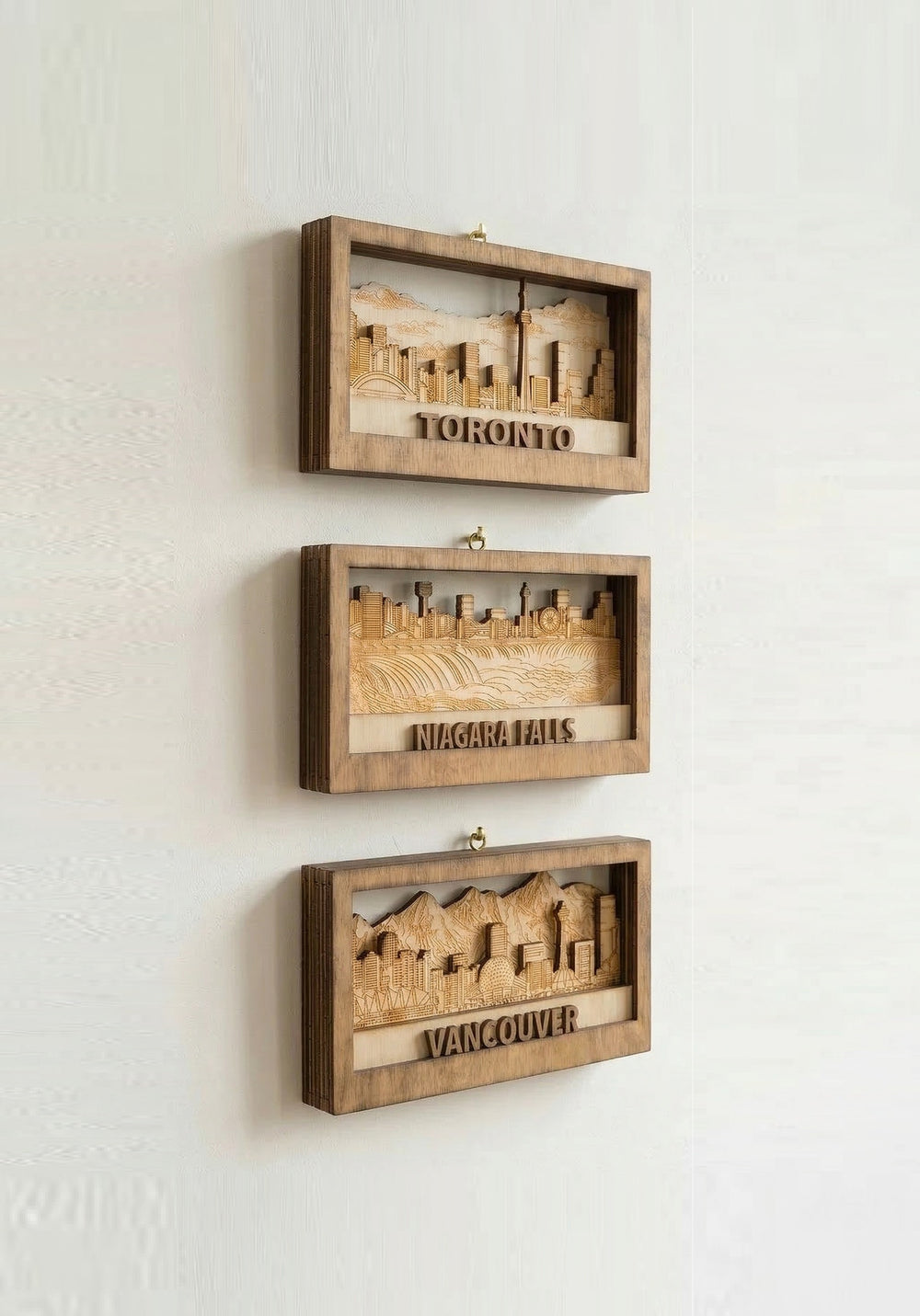 Three wooden wall art pieces displaying cityscapes of Toronto, Niagara Falls, and Vancouver on a white wall.